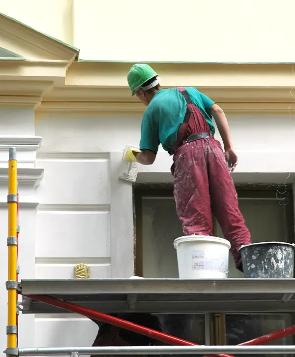 Painter on a ladder masking off trim at the peak of a house before spraying paint