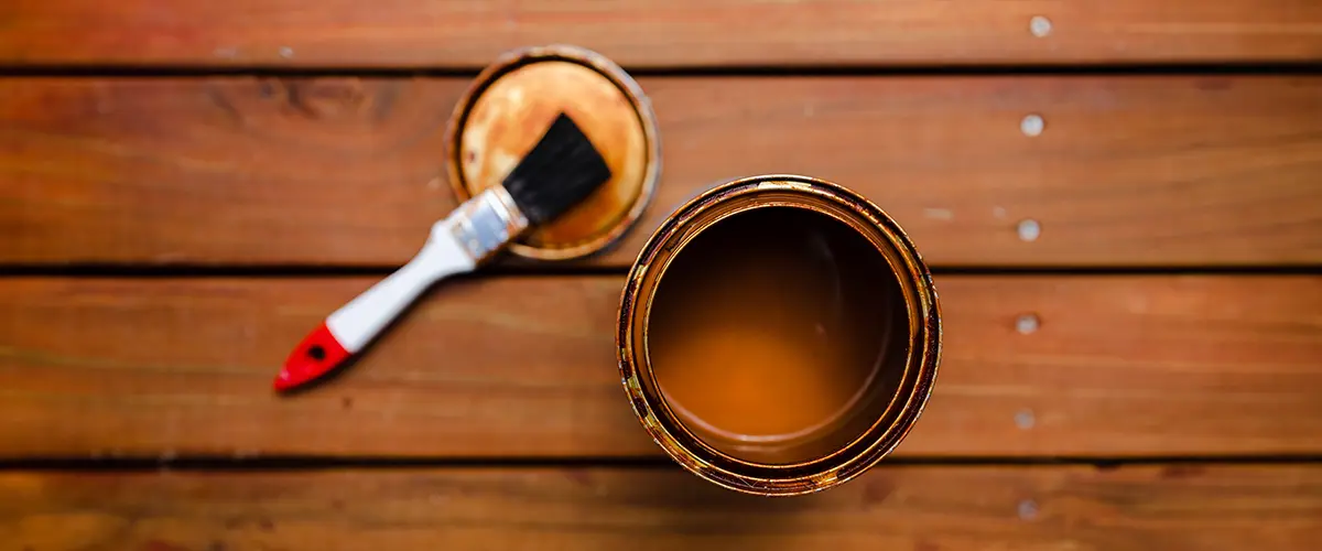 Open can of wood stain and paintbrush on a wooden deck, illustrating essential materials for DIY deck staining and maintenance.