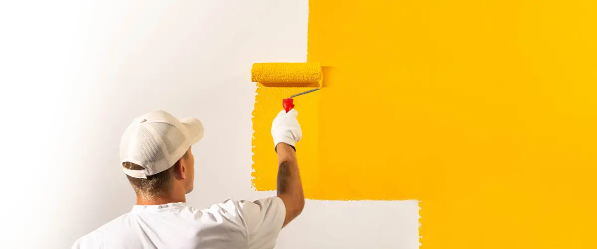 Professional painter in white outfit applying bright yellow paint on a wall with a roller, enhancing home interior aesthetics.