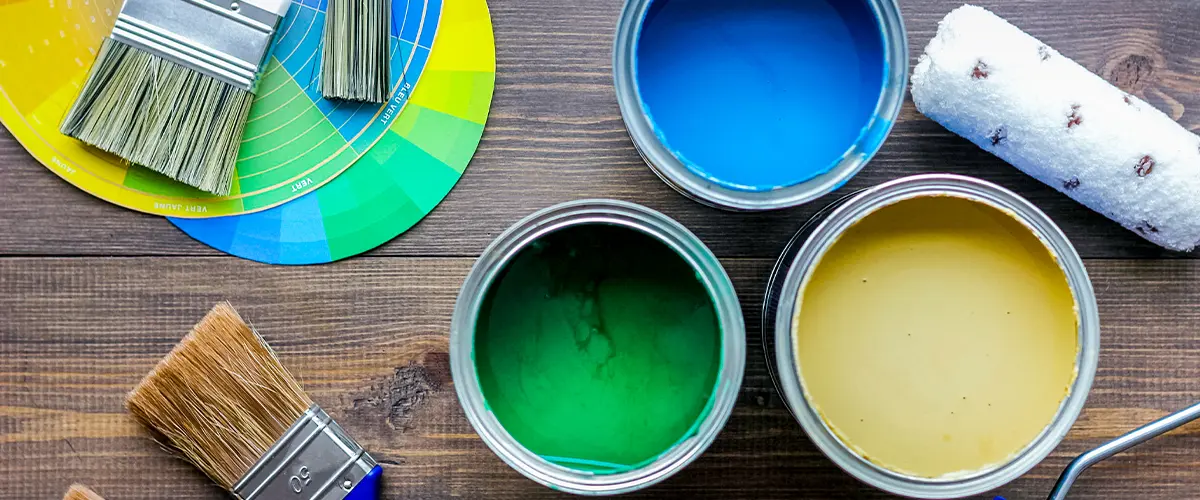 Open paint cans in blue, green, and yellow with brushes and color wheel for choosing the right shade. Oil vs Latex Exterior Paint