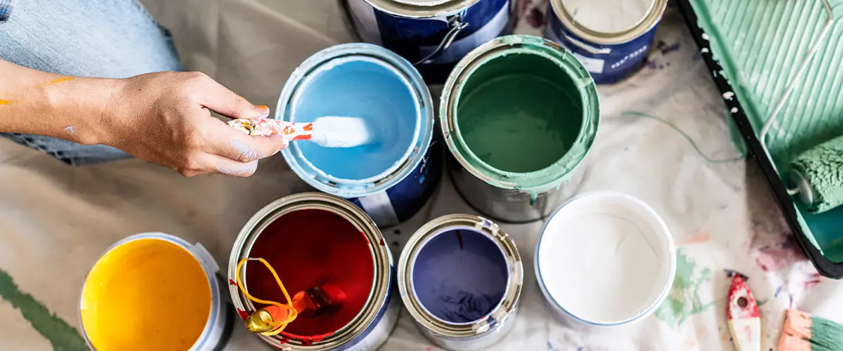 Person holding a paintbrush over various open paint cans for a home renovation project.