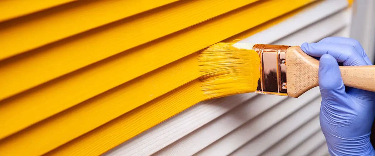 A person applying fresh yellow paint to exterior house siding with a paintbrush for a home improvement project.