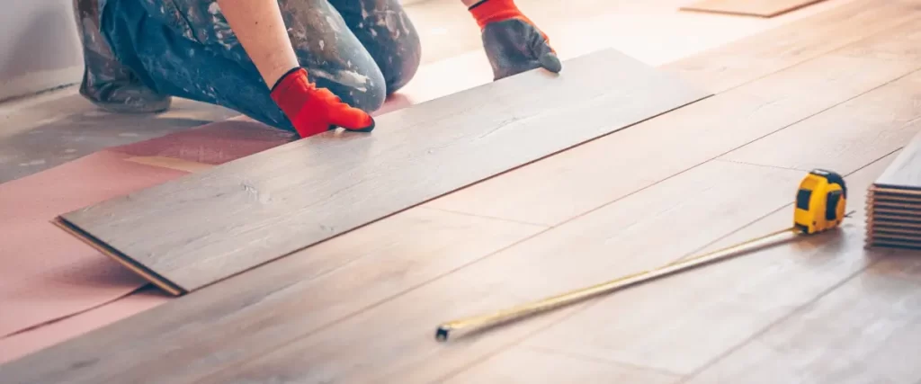 A person wearing red gloves installing light-colored wood flooring with a tape measure nearby