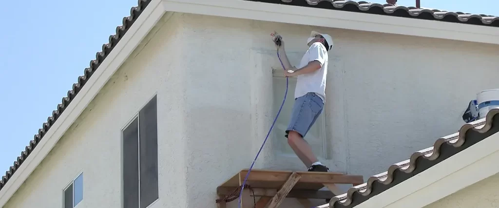 Painter spraying exterior wall on second floor of stucco house