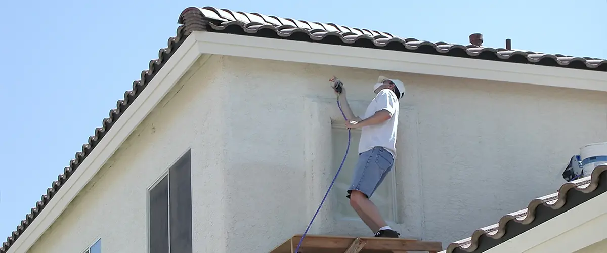 Painter using spray gun on upper stucco wall of two-story home