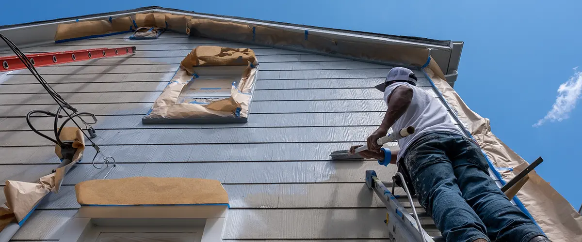 Painter on ladder applying coat to blue siding with windows masked