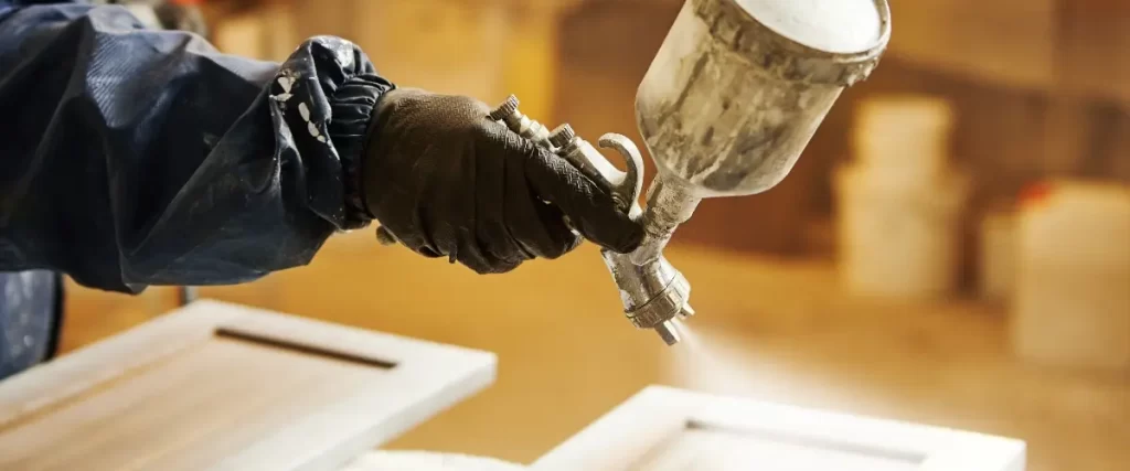 A person in a blue jacket and gloves sprays paint on a wooden surface