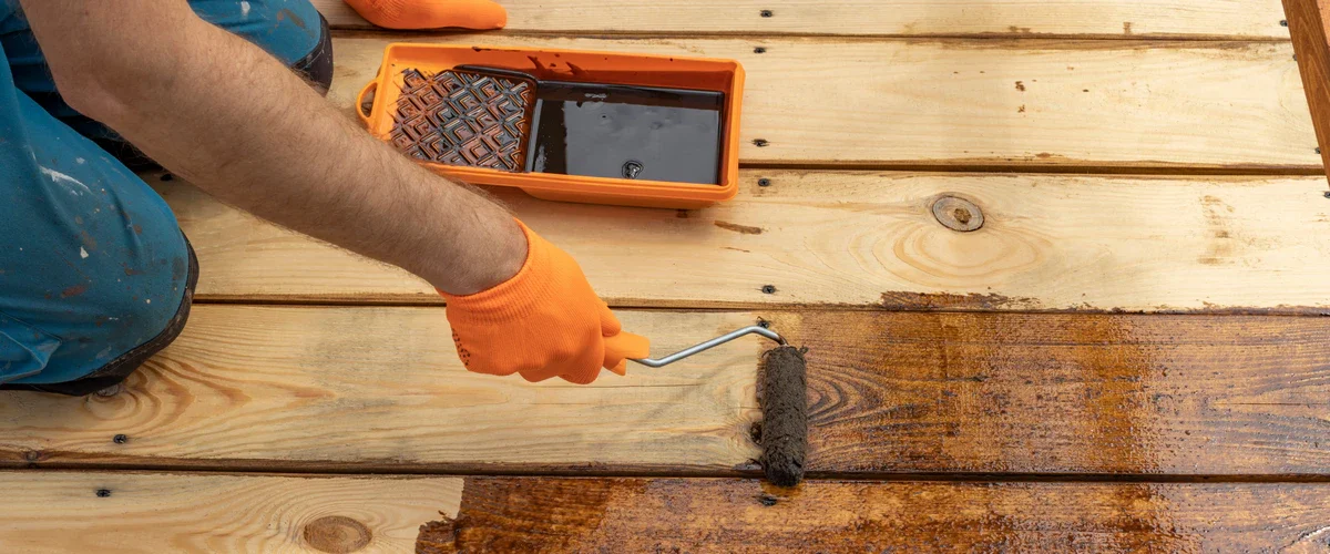 A close-up view of a person wearing orange gloves using a small roller to apply a dark brown protective staining finish to a light-colored wooden plank deck.