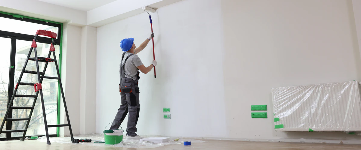 A professional painter wearing a blue hard hat and grey overalls uses a long-reach roller to apply fresh white house paint to a high interior wall in a modern room.