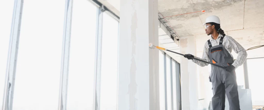 A professional painter wearing a white hard hat and grey overalls uses a long-handled roller to apply white paint to a concrete pillar inside a bright, modern commercial building with floor-to-ceiling windows.