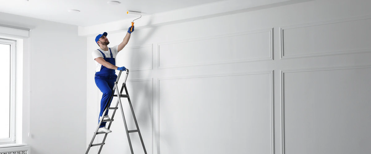 A professional male painter in blue overalls standing on a silver stepladder while using a roller to apply white paint to a modern interior wall featuring decorative panel molding.