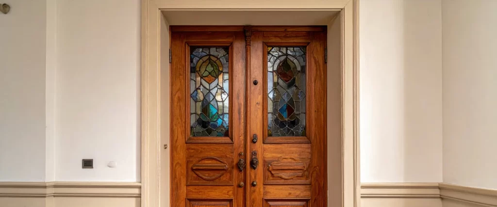 Detailed view of vintage carved wooden double doors featuring intricate stained glass inserts set against a clean creamy white entryway wall.