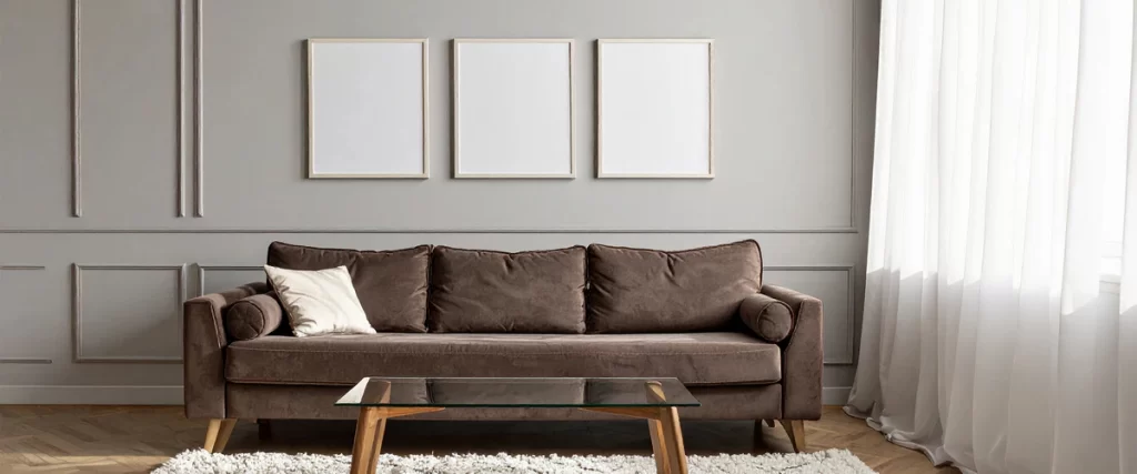 An elegant interior scene featuring cool gray walls with decorative panel molding, three empty frames, and a plush chocolate brown velvet sofa.