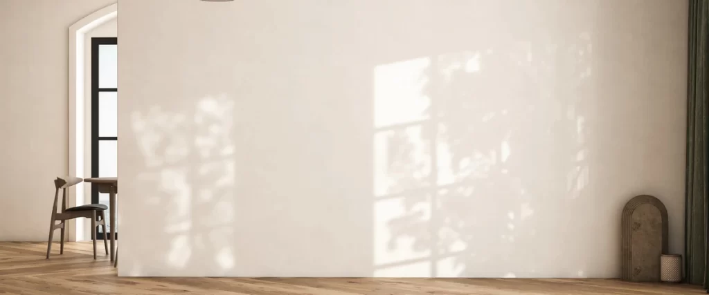 A minimalist interior space featuring a large blank wall painted in a warm white finish with natural sunlight and tree shadows cast across the surface.