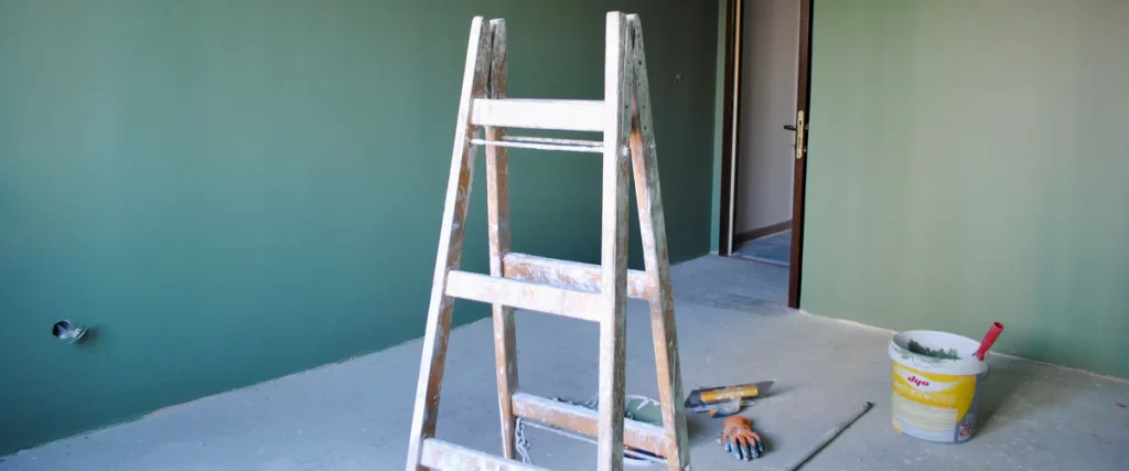 An interior room undergoing renovation with a wooden step ladder, paint buckets, and trowels used for preventing paint from cracking through proper prep.
