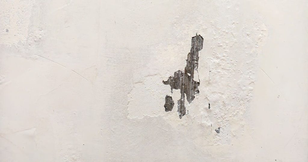 A close-up view of a white interior wall showing a large patch of peeling and cracking paint that has exposed the underlying surface.