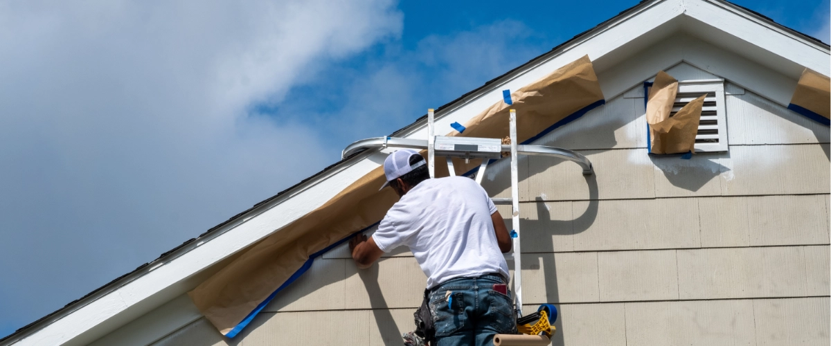 Exterior painter on a ladder spray painting house siding with windows masked off for protection.