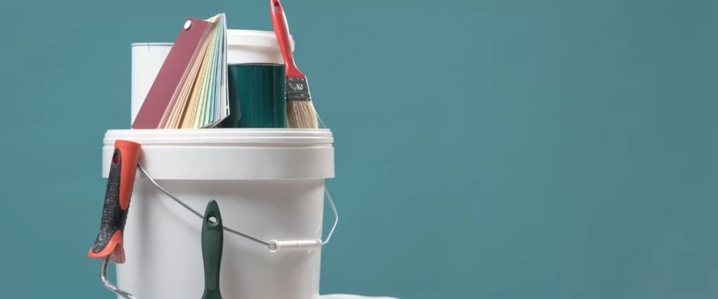 Paint bucket with brushes, roller, color swatches, and masking tape arranged on a table against a teal background.