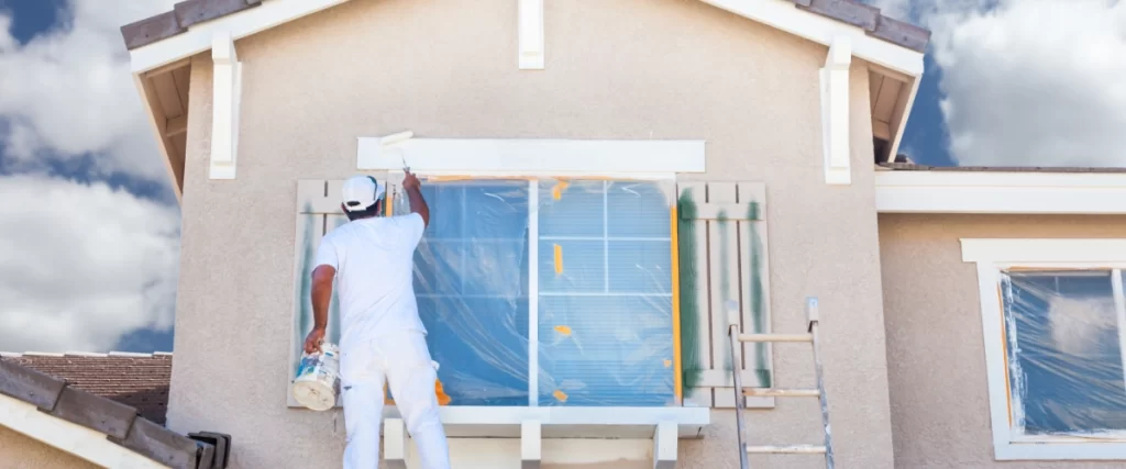 Professional house painter in white uniform painting exterior trim around a second-story window while standing on a roof.