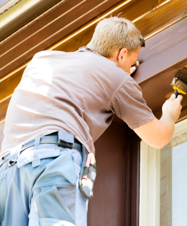 Contractor applying dark wood stain with a brush to exterior window trim on a modern wooden house.